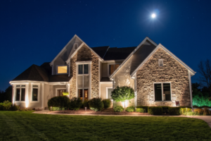 Dark Sky Compliant Outdoor Lighting - Elegant Outdoor Lighting Design Two-story suburban house at night with stone facade and warm interior lights, surrounded by landscaped shrubs and a lit lawn under a starry sky.