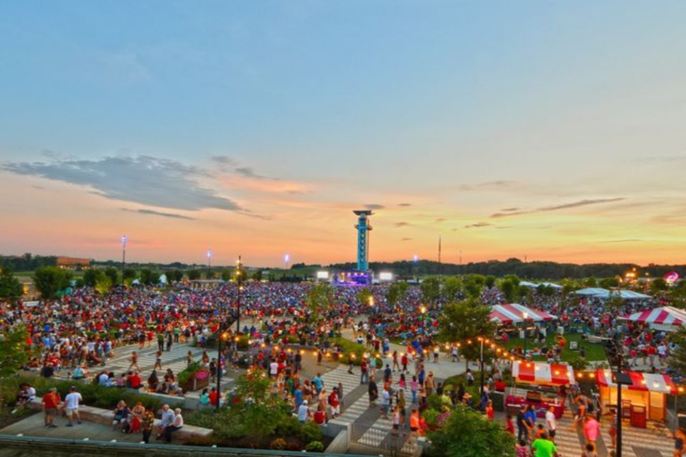 Blue Ash - Elegant Outdoor Lighting Design Wide view of a crowded outdoor festival at sunset with stalls, tents, and a distant stage.