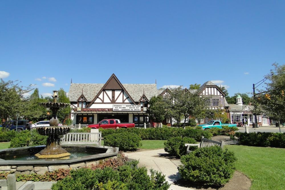 Mariemont - Elegant Outdoor Lighting Design Quaint Tudor-style storefronts line a sunny street with a tiered fountain in the foreground and trimmed hedges, benches nearby.