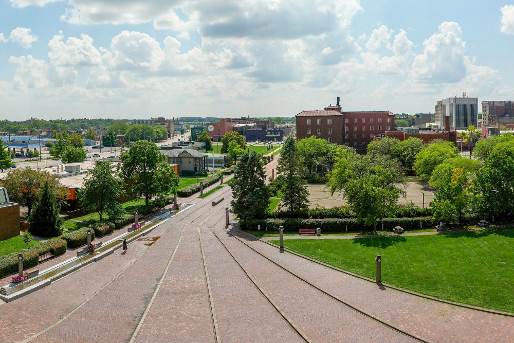 MiddletownOH - Elegant Outdoor Lighting Design Wide view of a brick-paved plaza with tram tracks converging toward a city block, flanked by green lawns and trees under a blue, cloudy sky.
