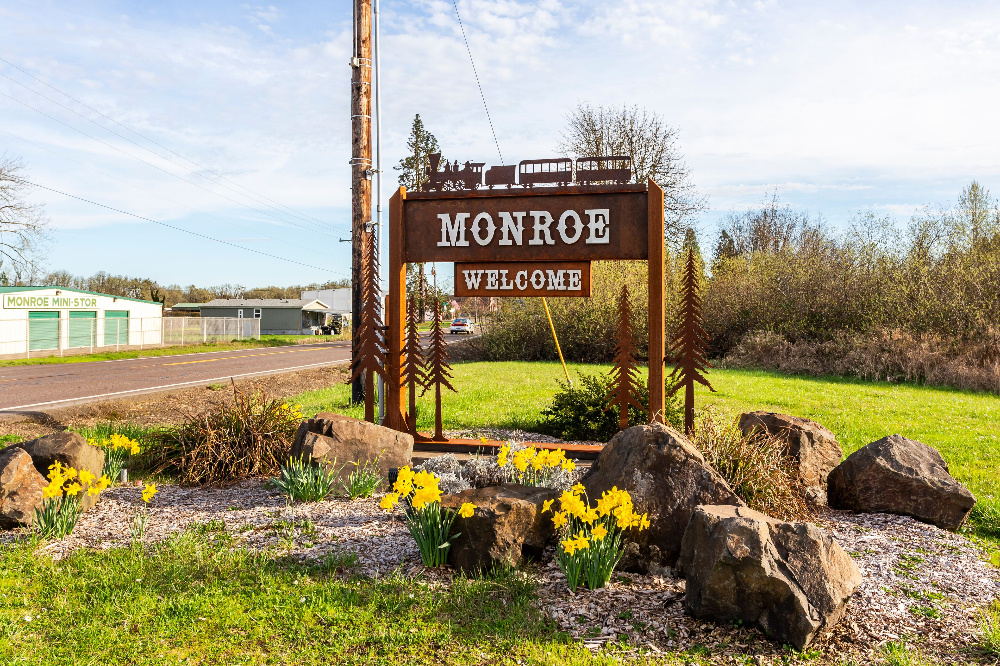 Monroe - Elegant Outdoor Lighting Design Monroe welcome sign with a train silhouette, set in a rock-lined garden with yellow daffodils and greenery nearby.